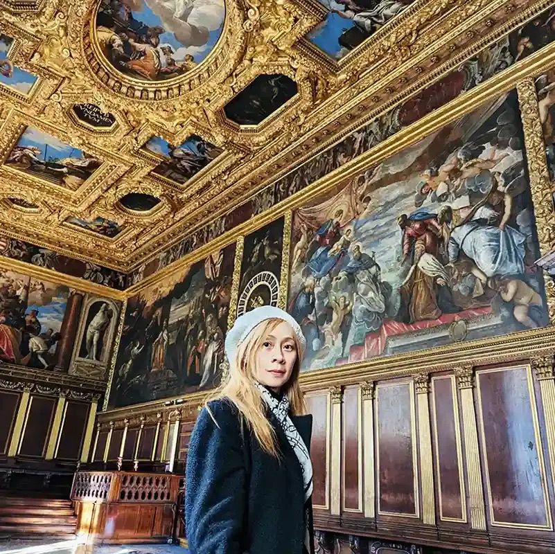 Montessori art director and teacher Yuming Tseng stands inside a grand museum hall surrounded by historic Renaissance wall murals and an ornate gold ceiling.