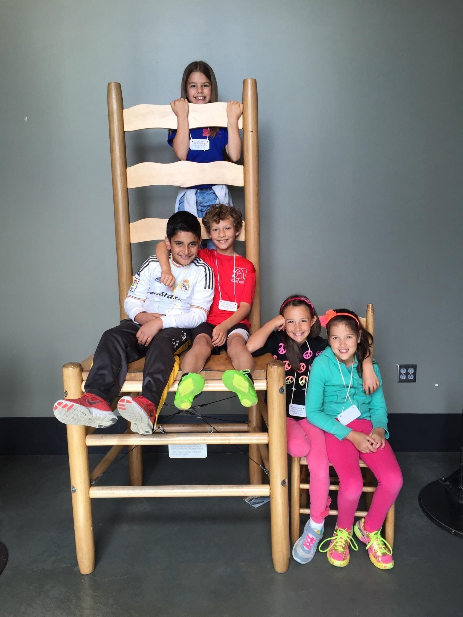 A group of smiling Montessori elementary students pose together on and next to a giant wooden oversized chair exhibit during a school field trip.