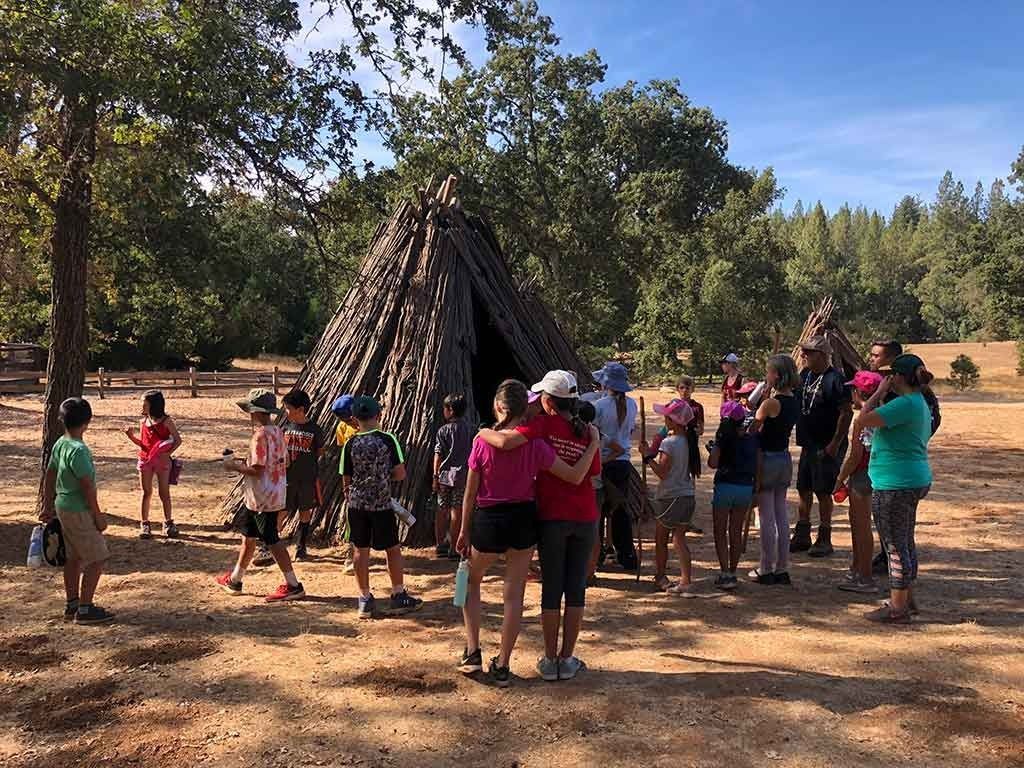 A group of Montessori elementary students and guides gather around a traditional Indigenous bark-covered teepee structure during an outdoor history field trip.
