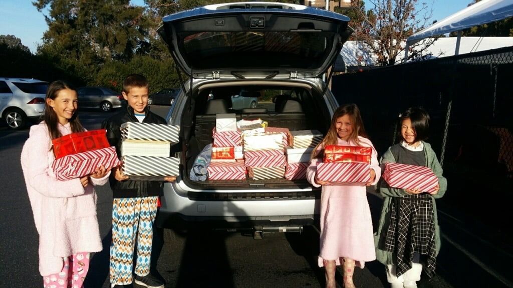 Four smiling Montessori elementary students hold stacks of gift-wrapped boxes while standing behind a car trunk loaded with donations for a holiday charity drive.