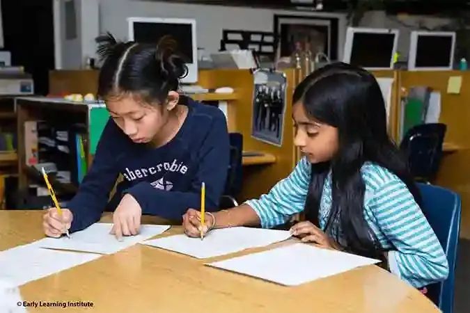 Two upper elementary Montessori students work collaboratively at a wooden table, discussing their writing assignments and pointing to their papers with pencils.