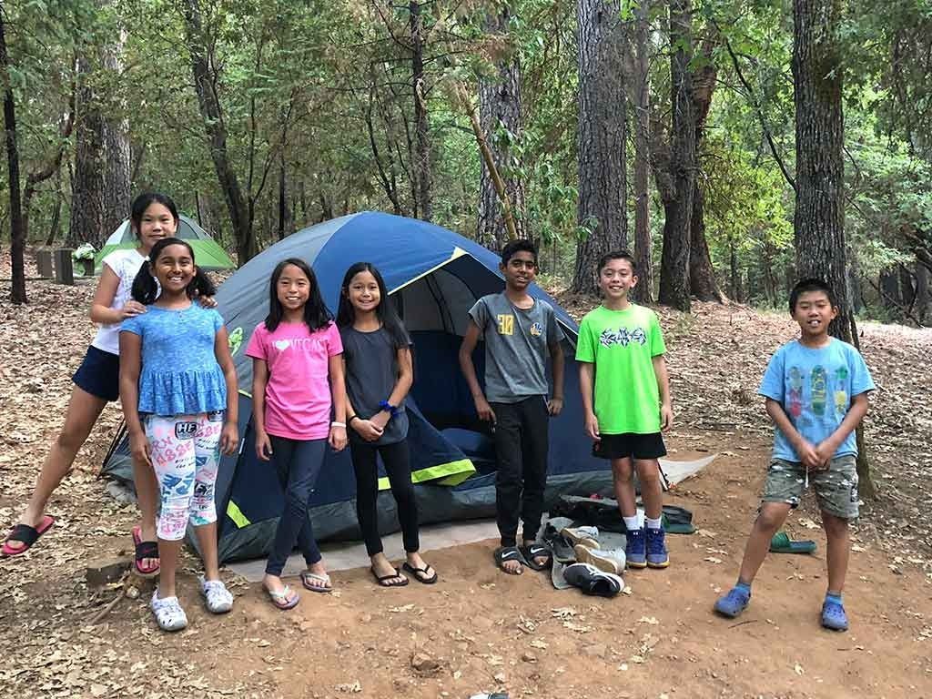 A group of smiling elementary students poses together outside a blue camping tent pitched in a wooded forest during an outdoor education field trip.