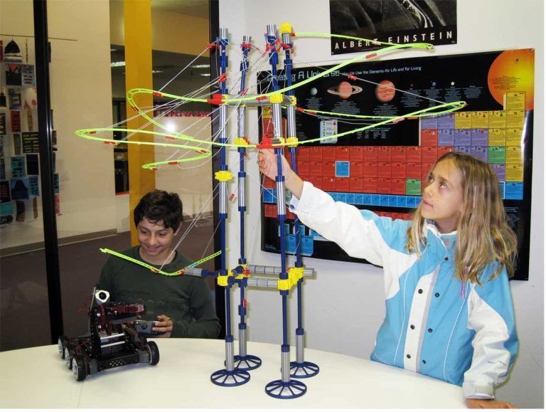 Two Montessori middle school students work on a complex STEM engineering project, assembling a multi-tiered marble run track structure alongside a programmable robotics vehicle.