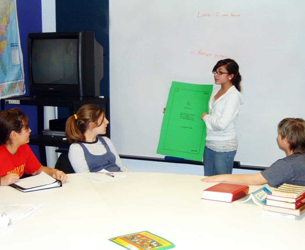 A Montessori middle school student presents a green poster project to classmates gathered around a table for a collaborative seminar discussion.