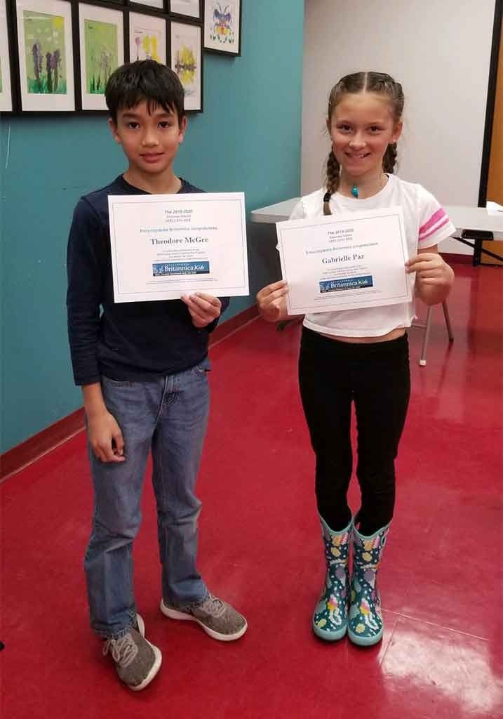 Two elementary students smile while holding up academic achievement certificates.
