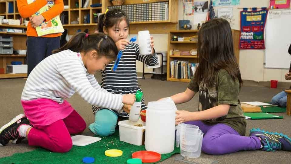 Three preschool students work collaboratively on a classroom floor mat, using scoops, measuring cups, and jars to practice pouring and volume concepts.
