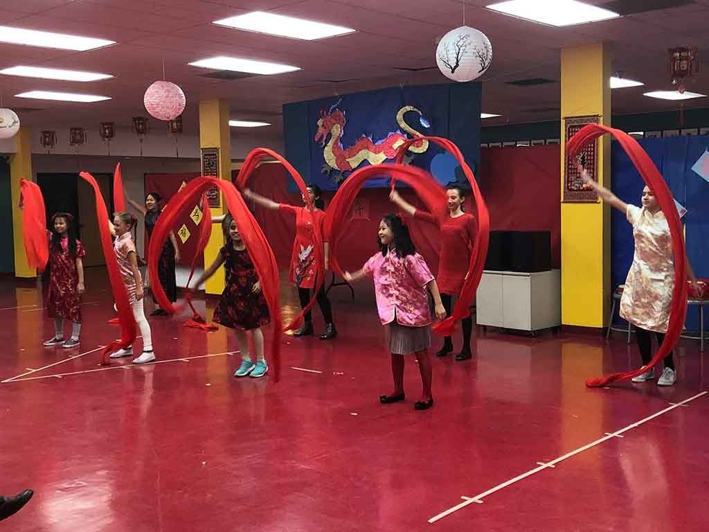 A group of elementary students perform a traditional Chinese ribbon dance with long, flowing red ribbons during a school cultural stage celebration.