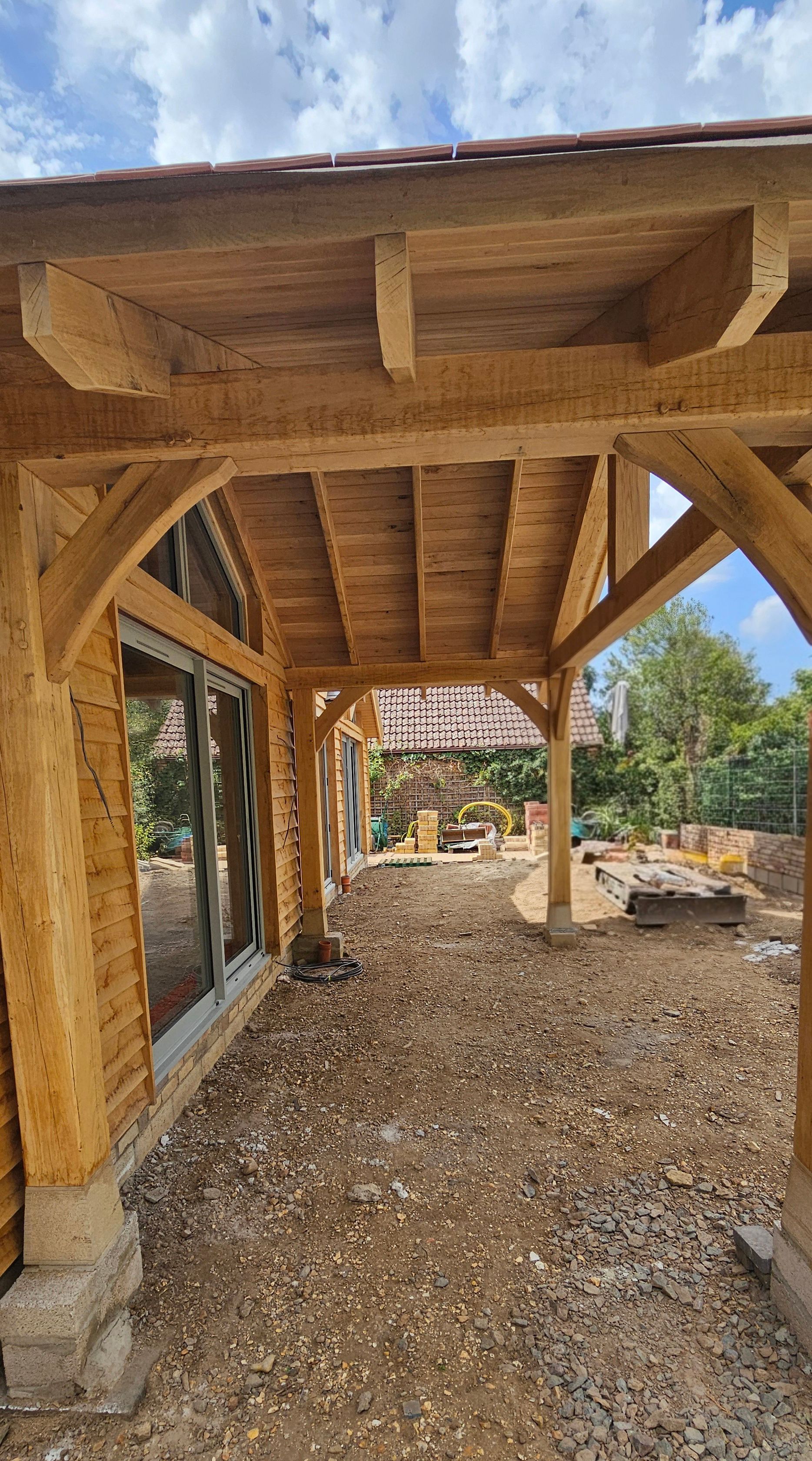 Oak porch structure under construction with exposed beams, stone pillar bases, and a gravel ground surface.