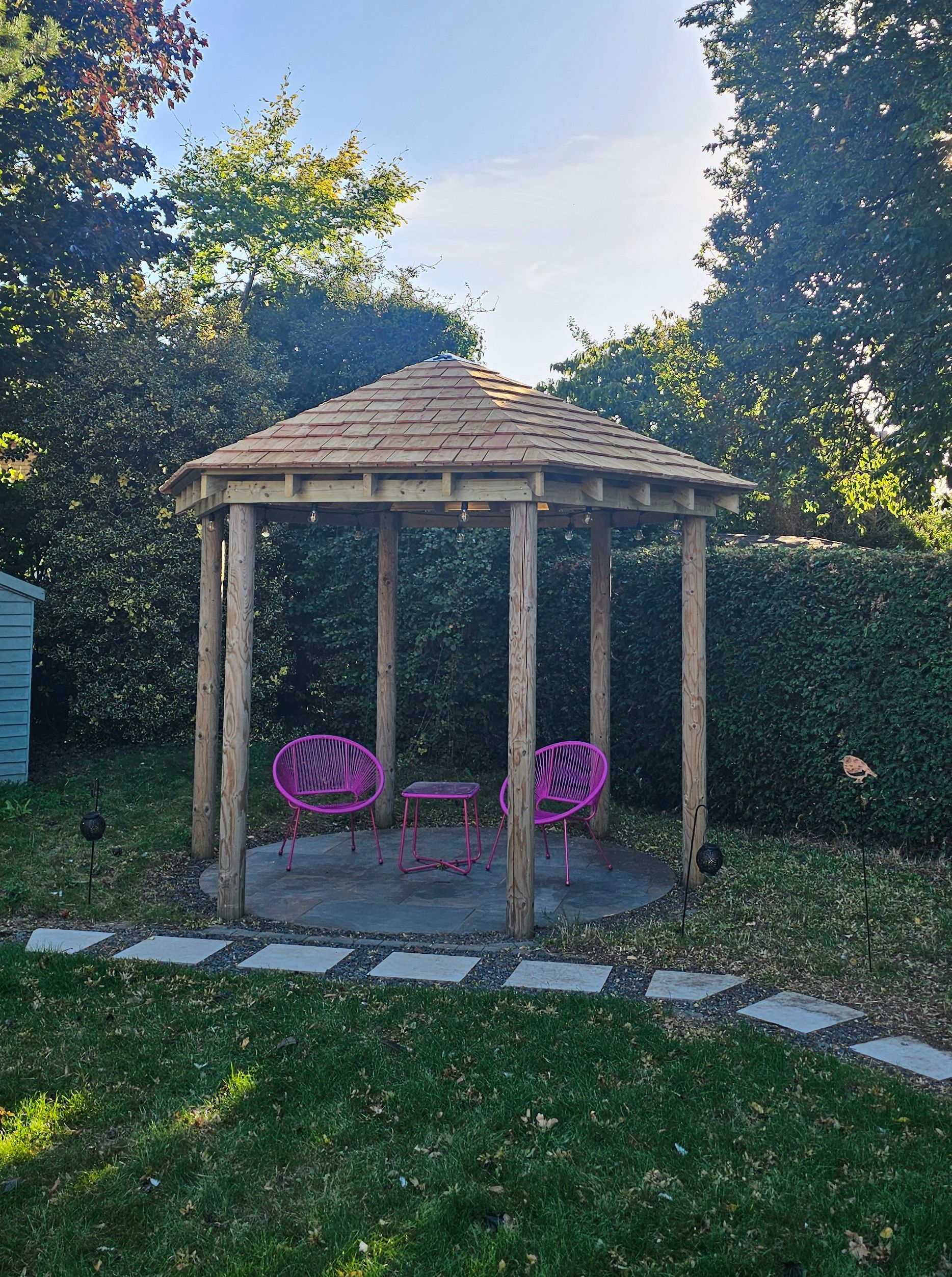 Oak wooden gazebo stands in a garden with two bright pink chairs and a small table on a stone floor, bordered by hedges.