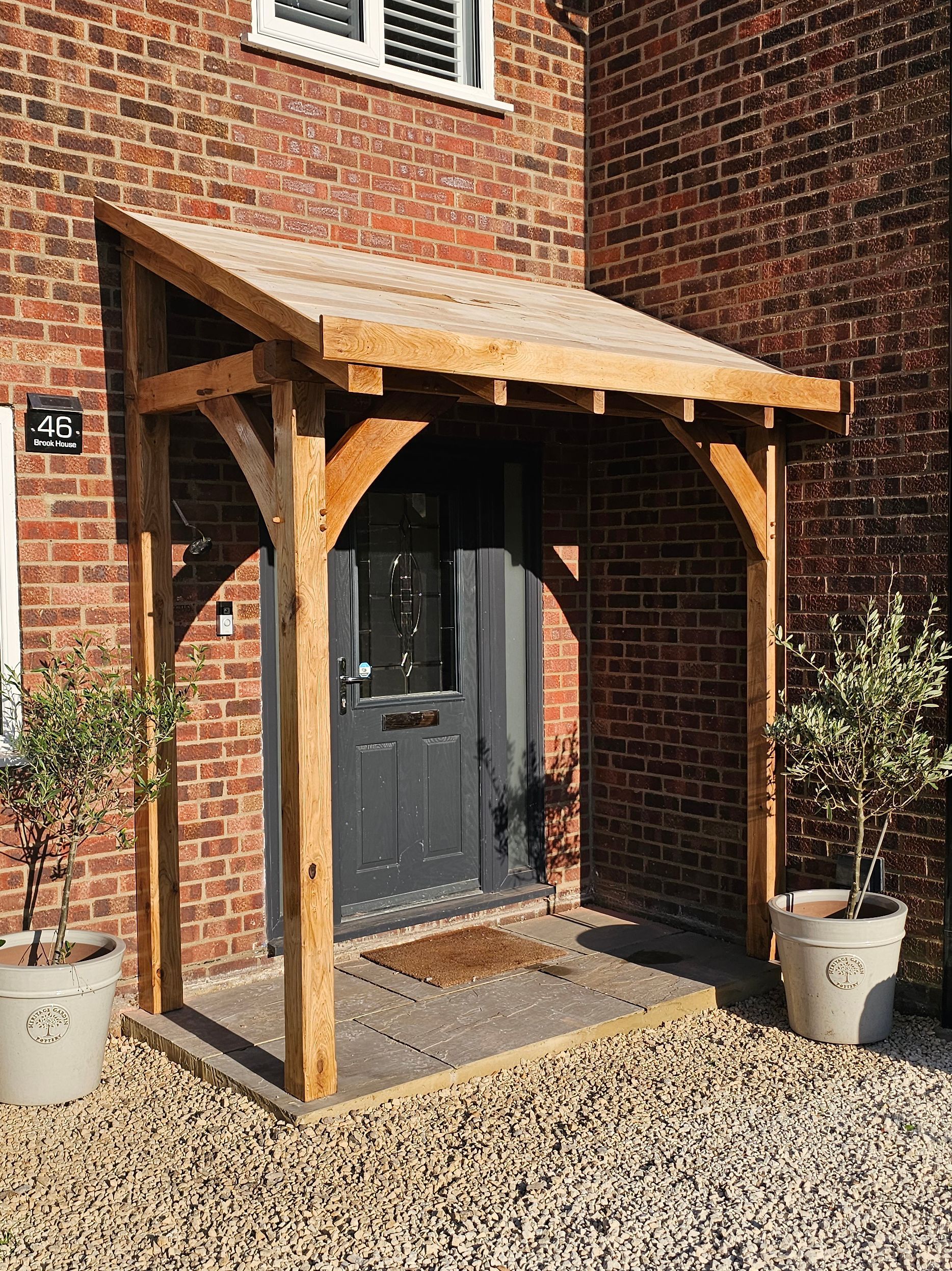A oak porch canopy with curved braces over a dark grey front door on a brick house, flanked by two potted olive trees.