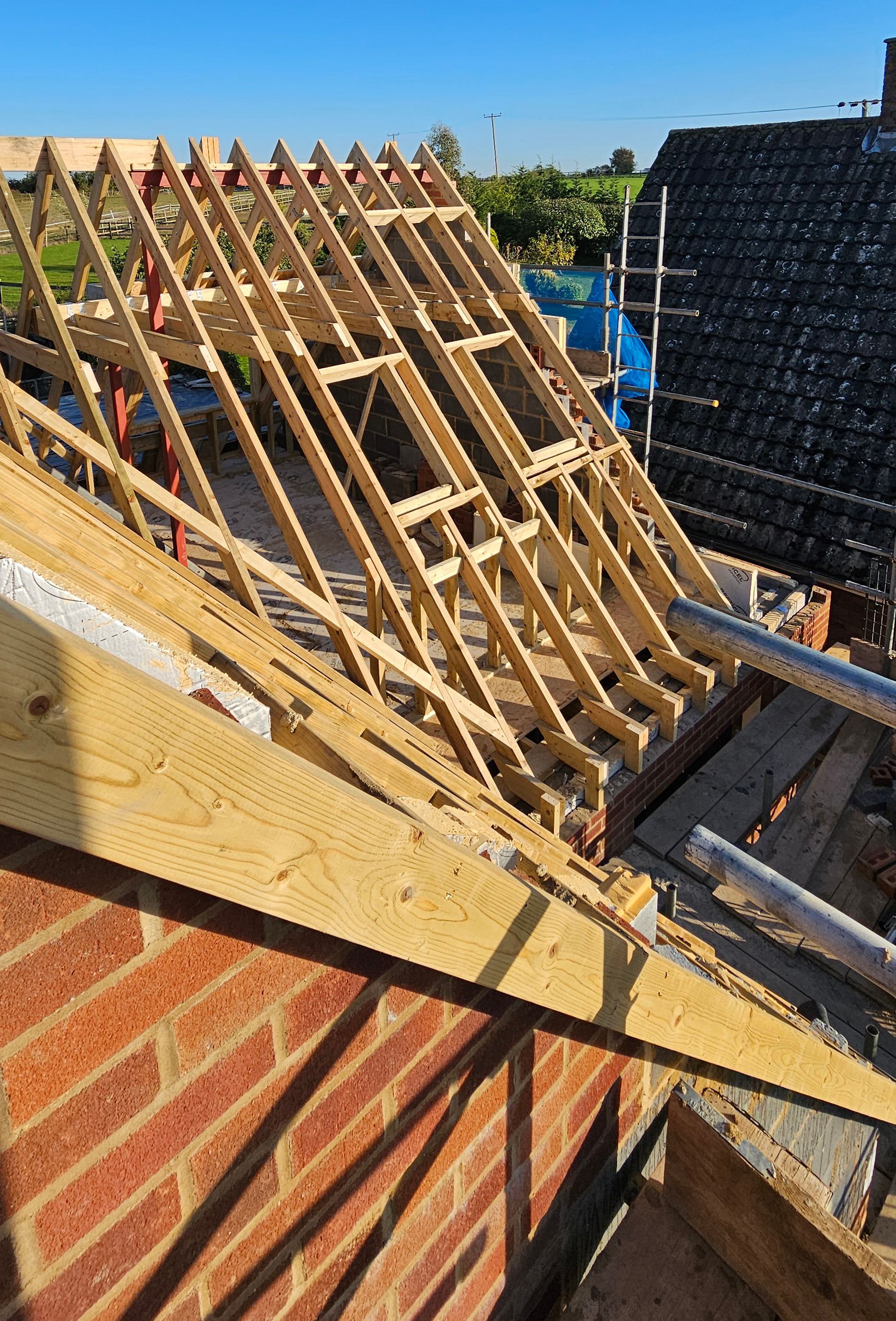 Construction view of a hand cut wood-framed house roof with a partially built dormer window, seen from scaffolding.