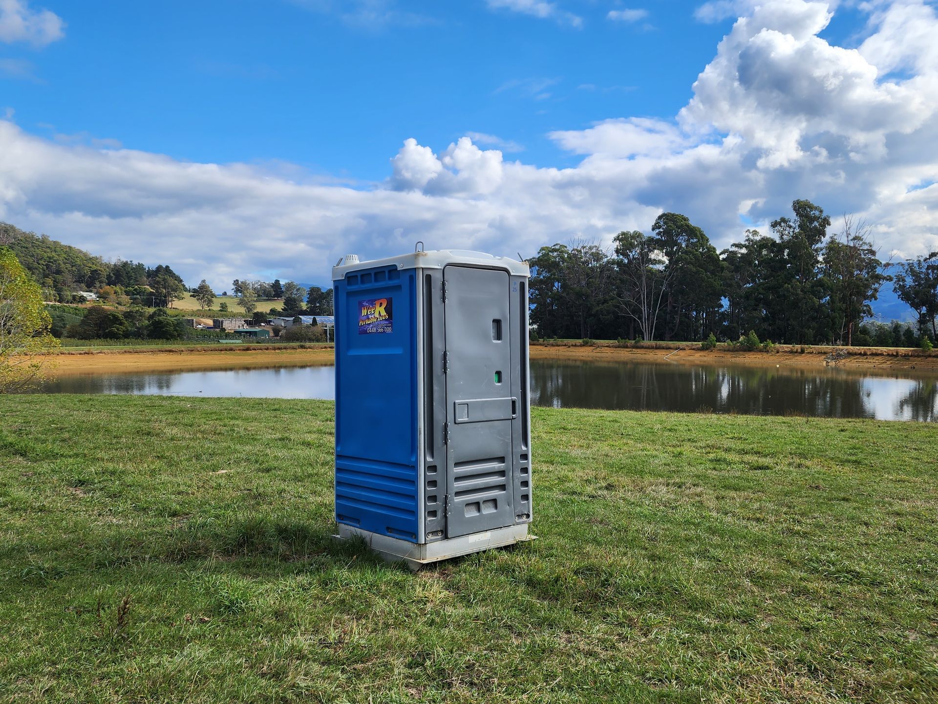 Blue portable toilet on grassy field beside a lake under a cloudy sky — Wee R Portable Loos in Glenorchy, TAS