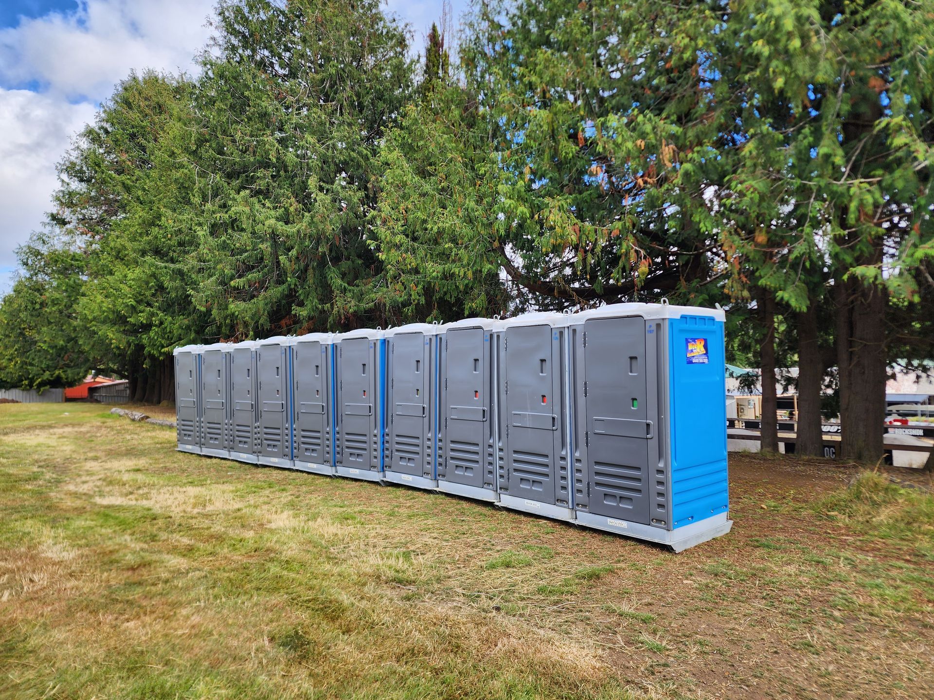 Row of portable toilets on grass beside trees, including a bright blue unit at the end  Wee R Portable Loos in Glenorchy, TAS