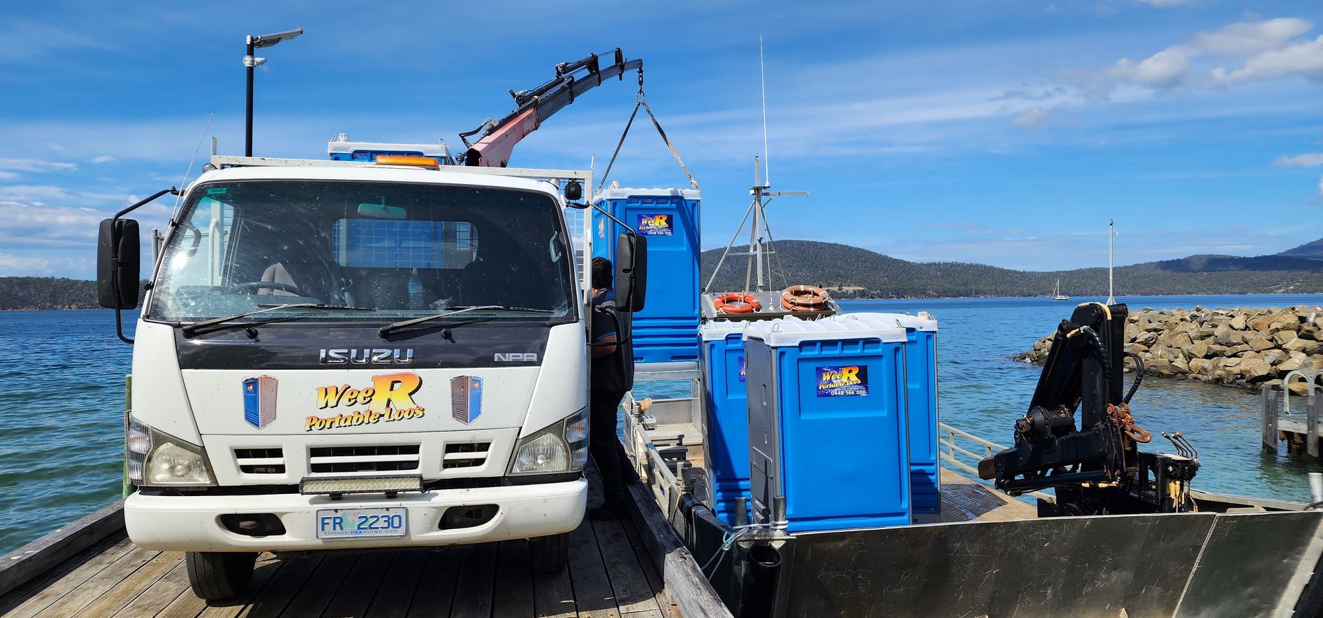 White utility truck parked on a pier beside blue equipment and a crane, with water and hills in the background - Wee R Portable Loos in Glenorchy, TAS