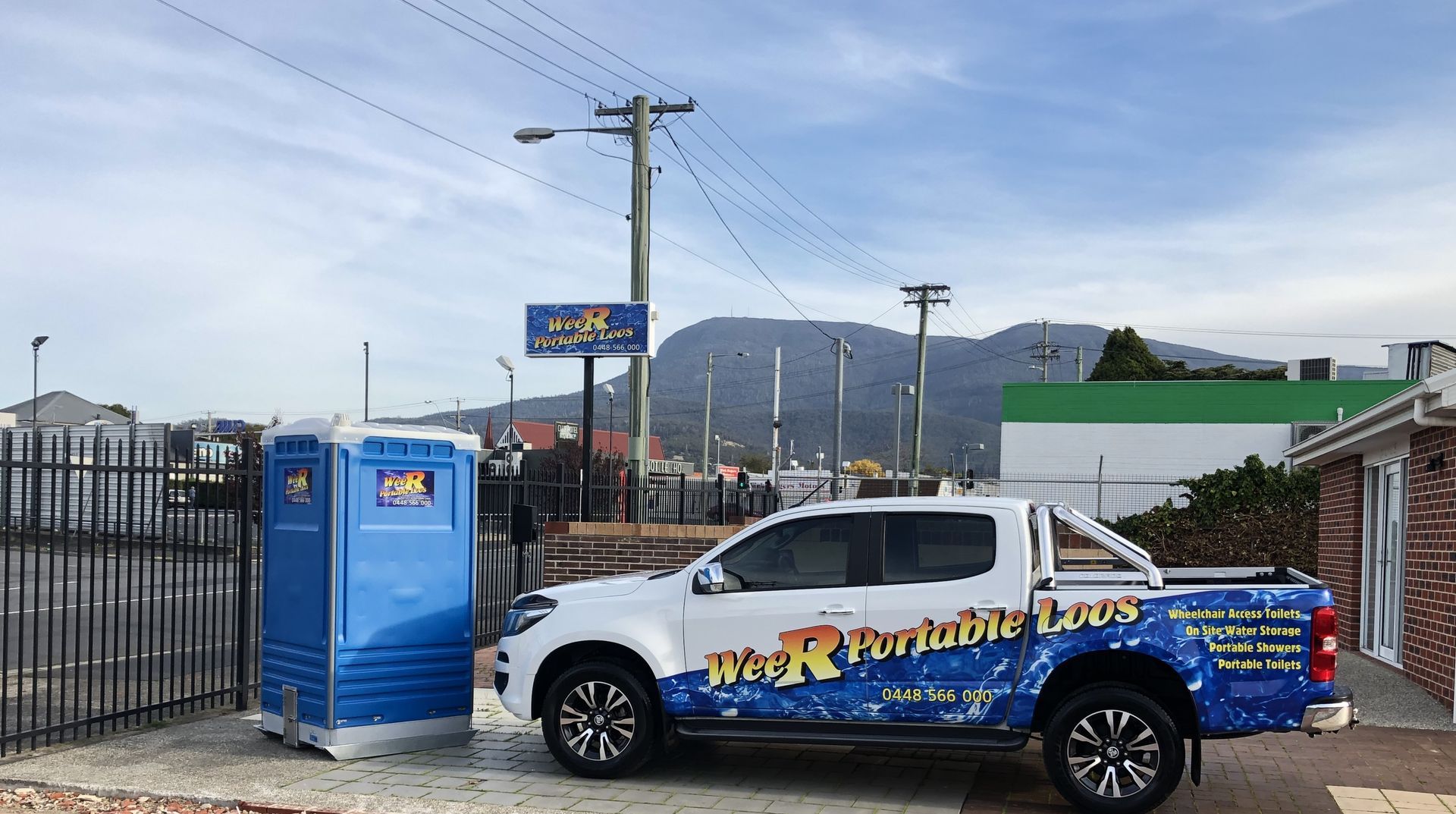 Blue pickup truck with racing graphics parked beside a portable toilet and black fence under a cloudy sky - Wee R Portable Loos in Glenorchy, TAS