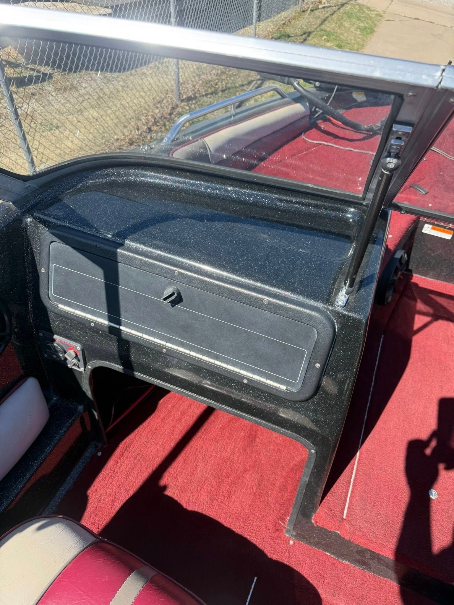 Black metal storage box with a latch on a red carpeted vehicle interior beside a mesh window