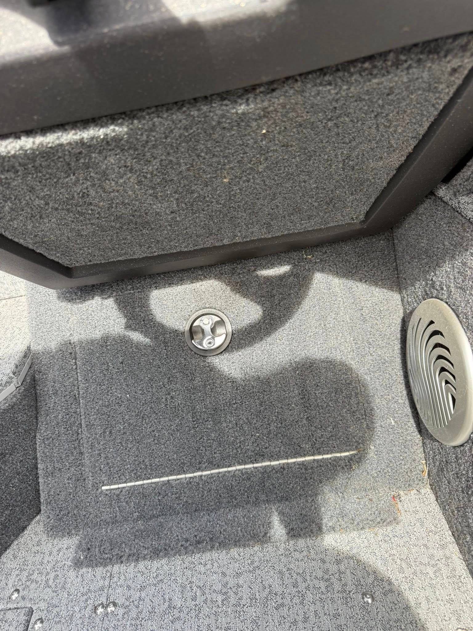 Gray carpeted compartment with a small round fitting, a rectangular floor hatch, and a vent on the right.