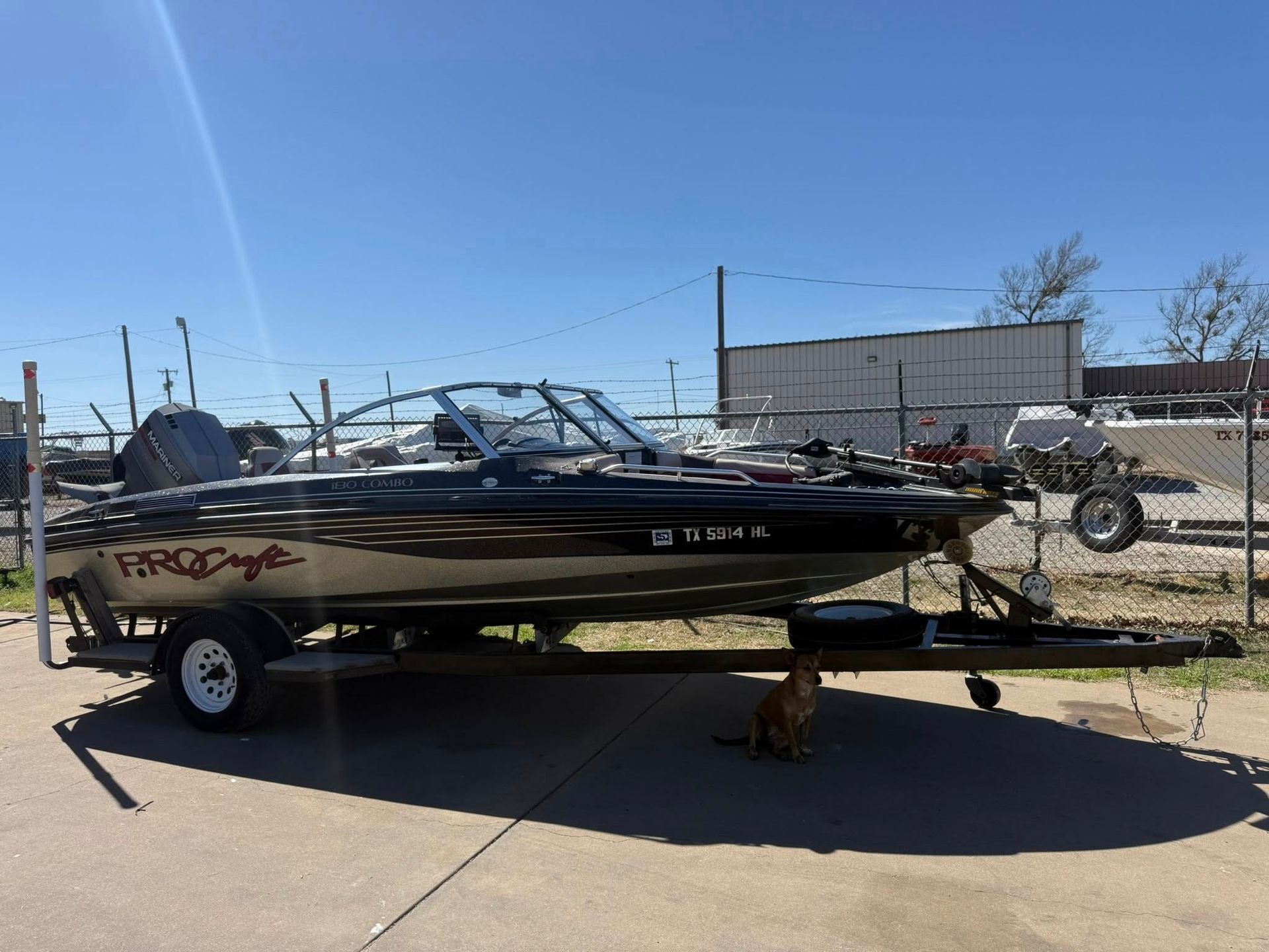 Black bass boat on a trailer in a sunny parking lot, with a dog underneath the hitch.
