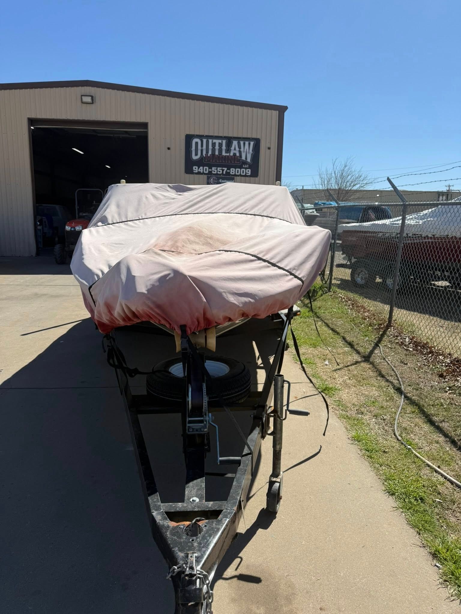 Boat on a trailer under a cover outside a garage, with a sign on the building and a fence nearby