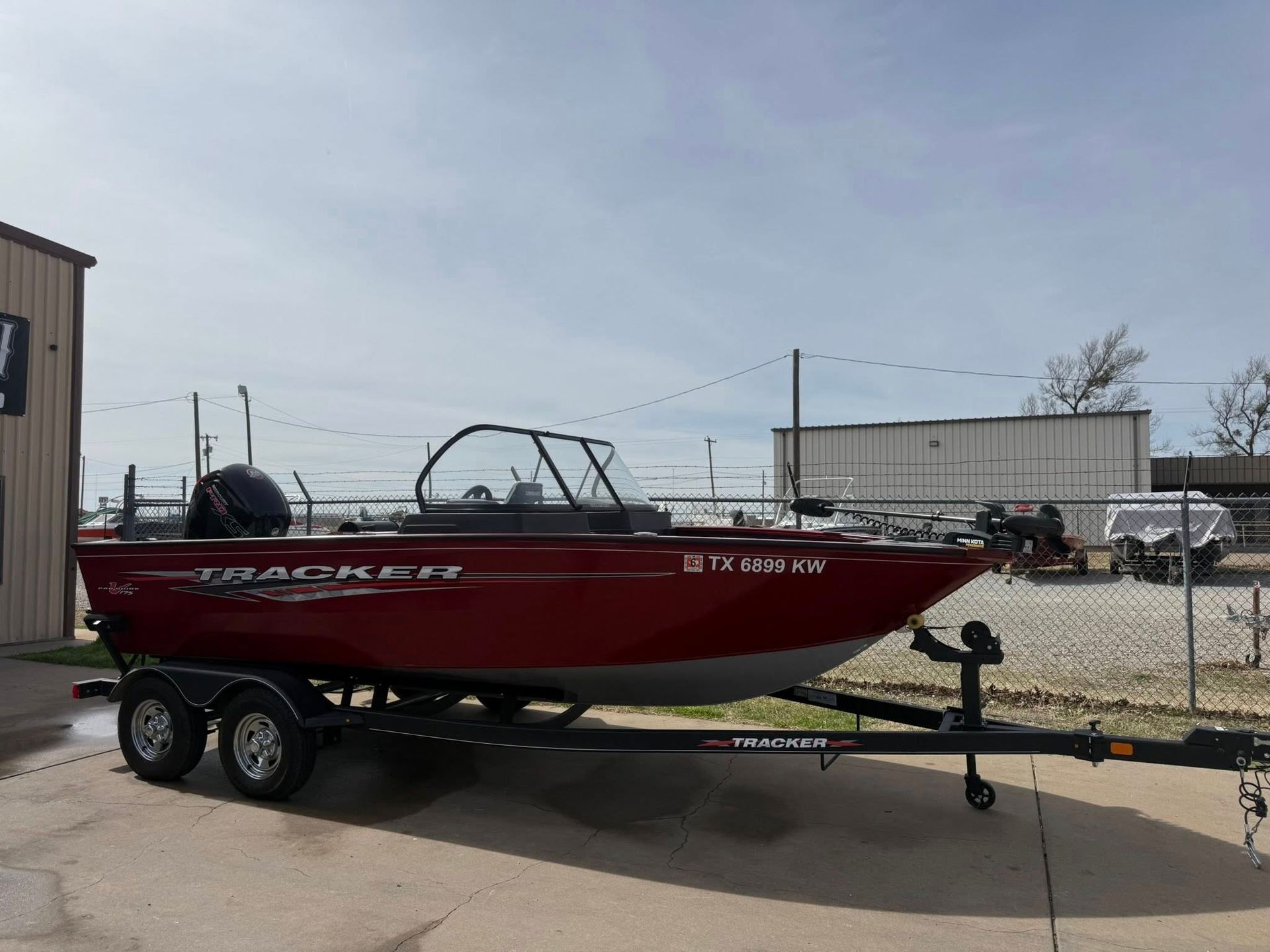 Red fishing boat on a trailer parked outdoors beside a building and chain-link fence.