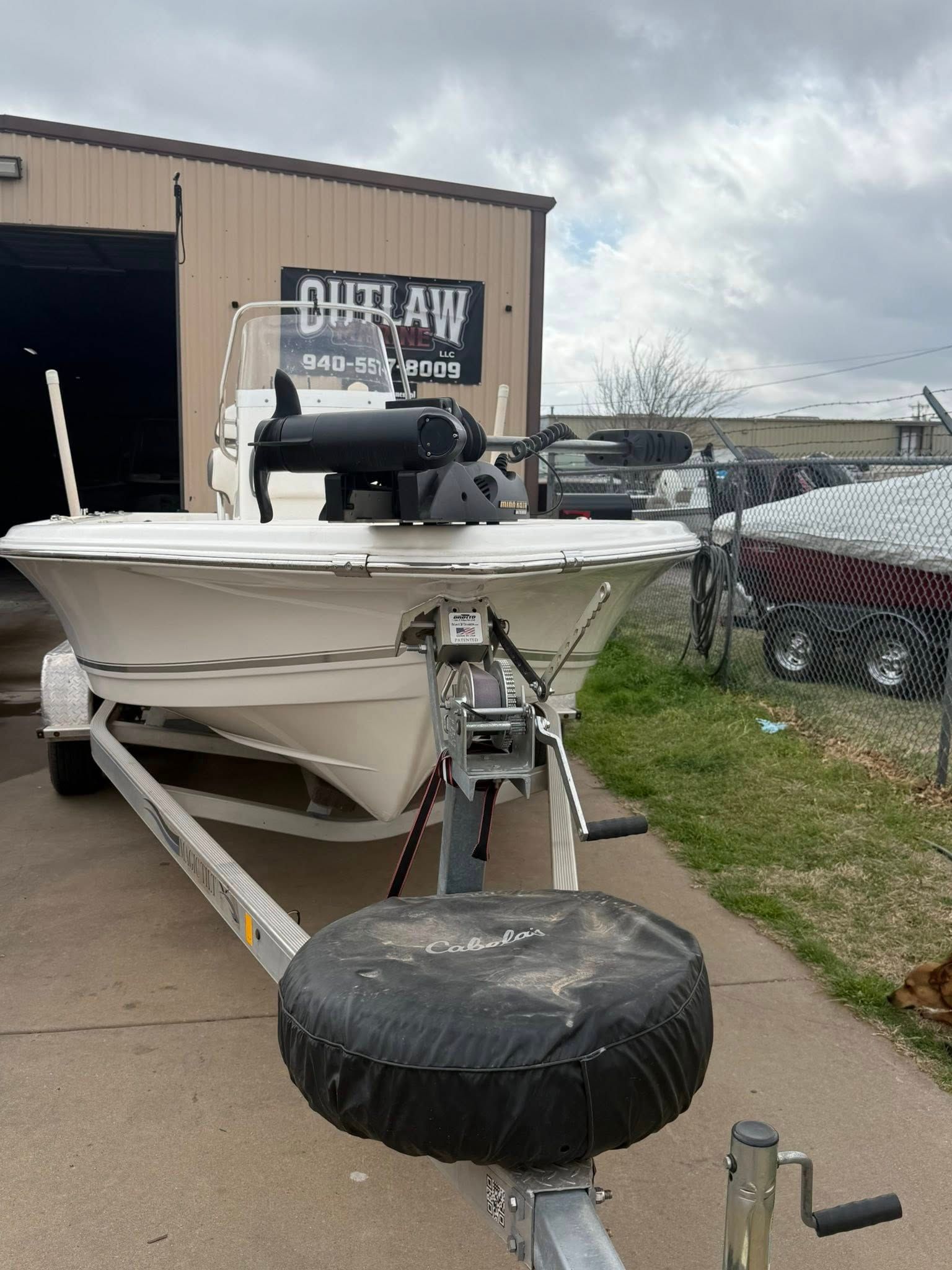 White motorboat on a trailer with a large black spare tire in front, parked by a garage and chain-link fence.
