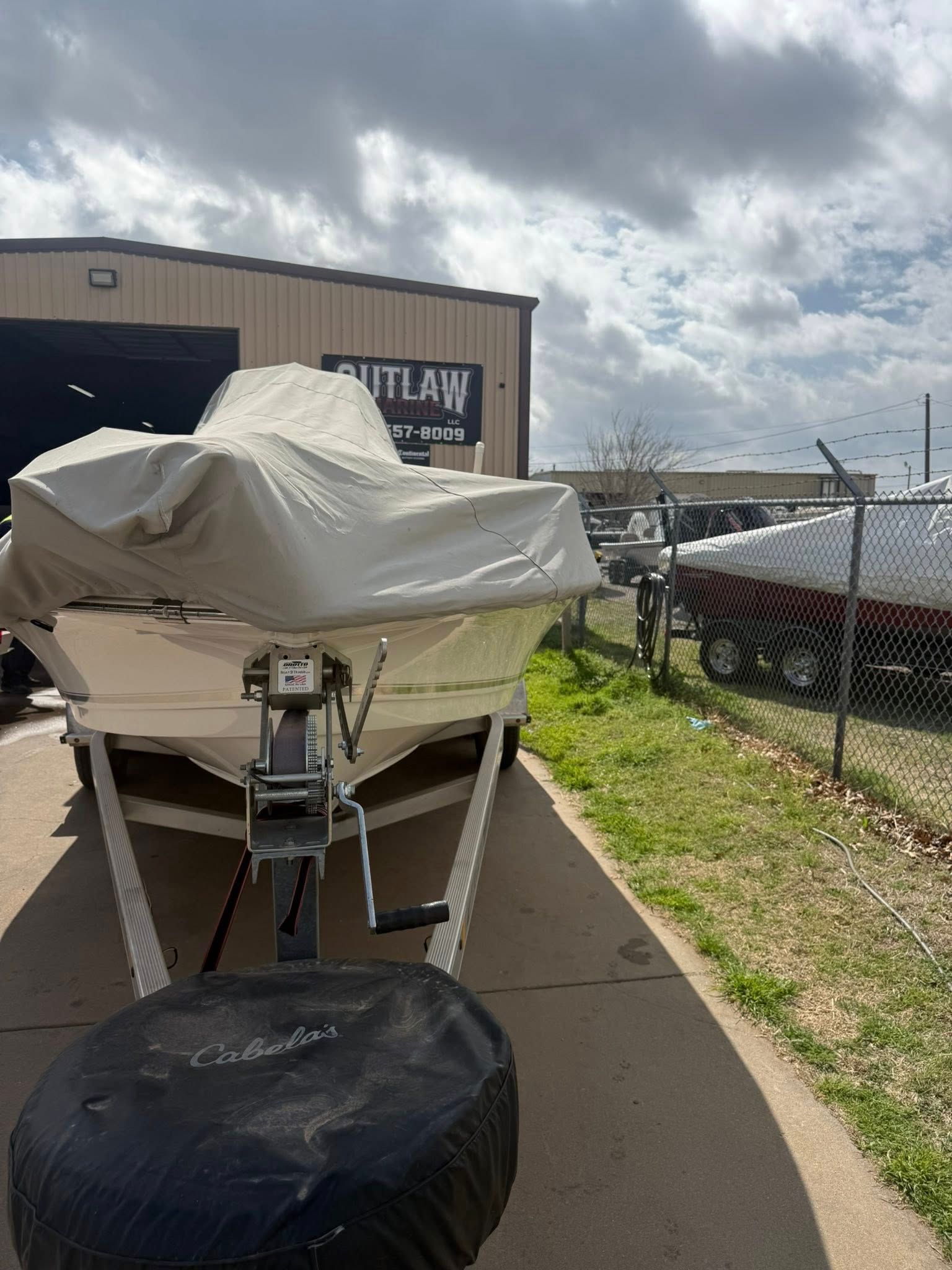 Boat on trailer covered with tarp beside a fence and building under cloudy sky