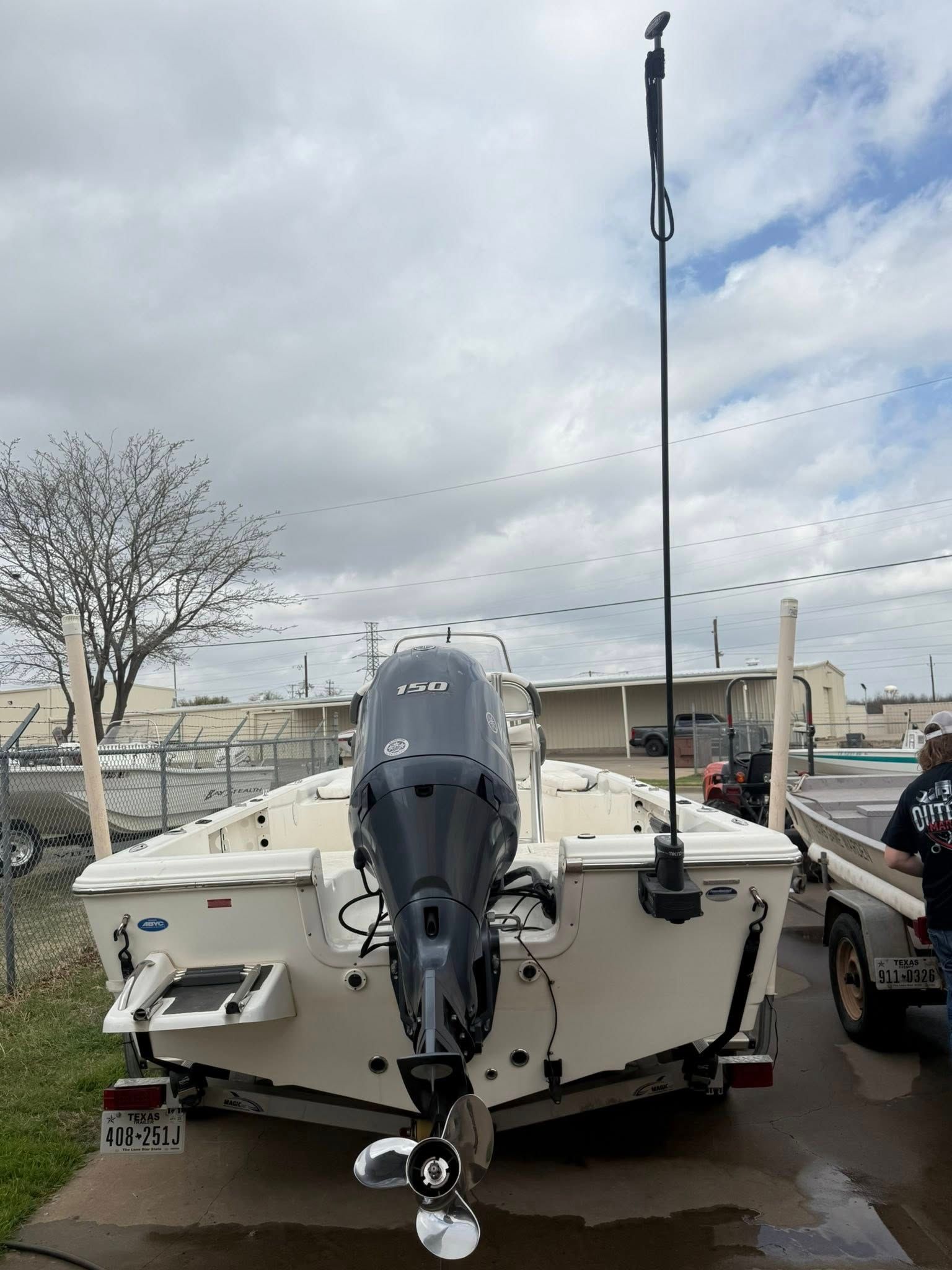 White boat with outboard motor and tall antenna parked on a trailer near a truck under cloudy skies