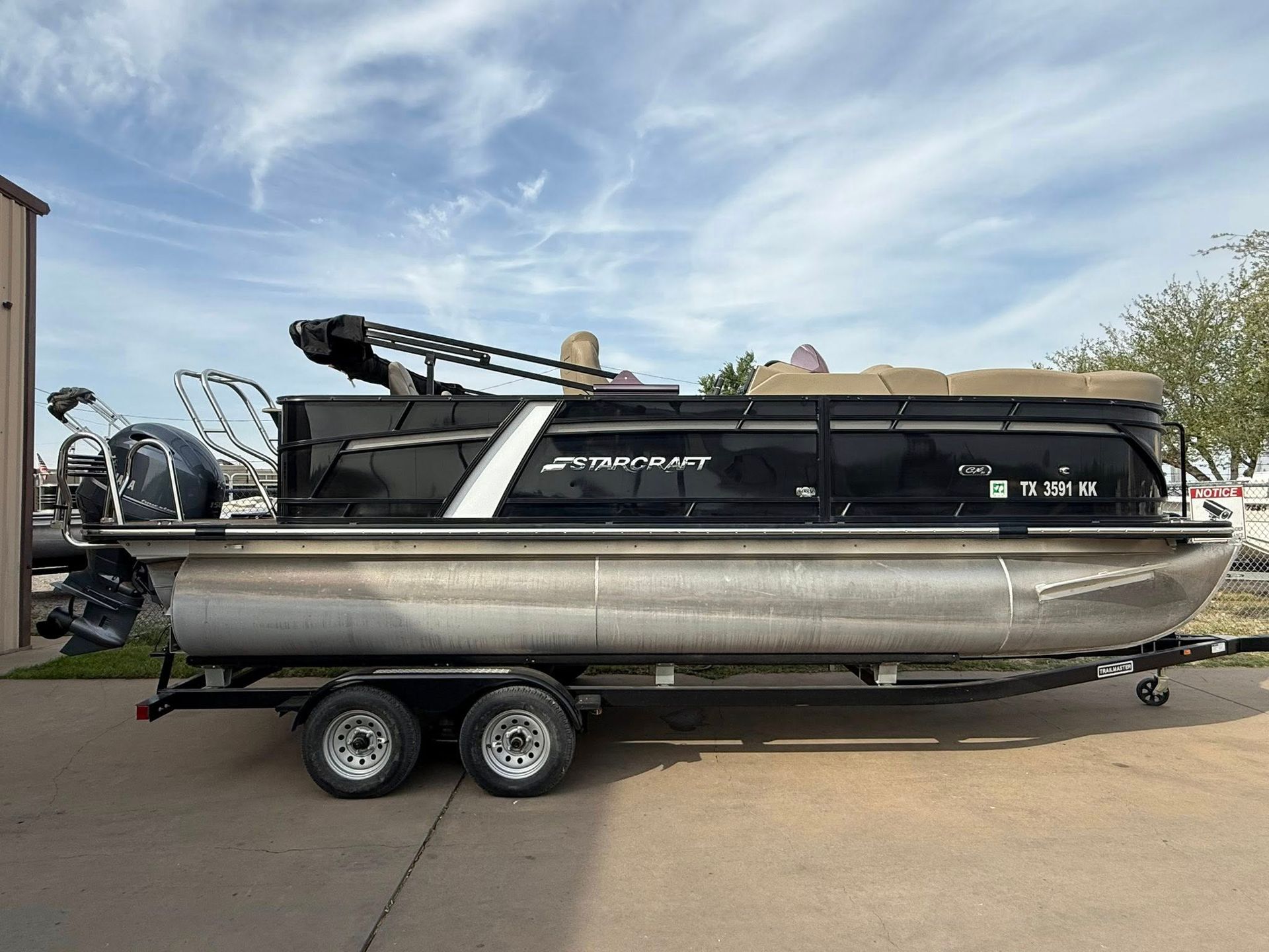 Black pontoon boat on a trailer parked outdoors under a cloudy sky.