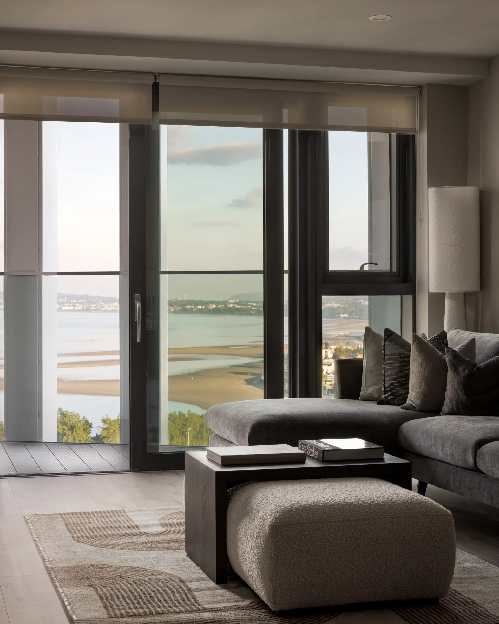 Modern living room with gray sofa, ottoman, and floor-to-ceiling windows overlooking a waterfront view
