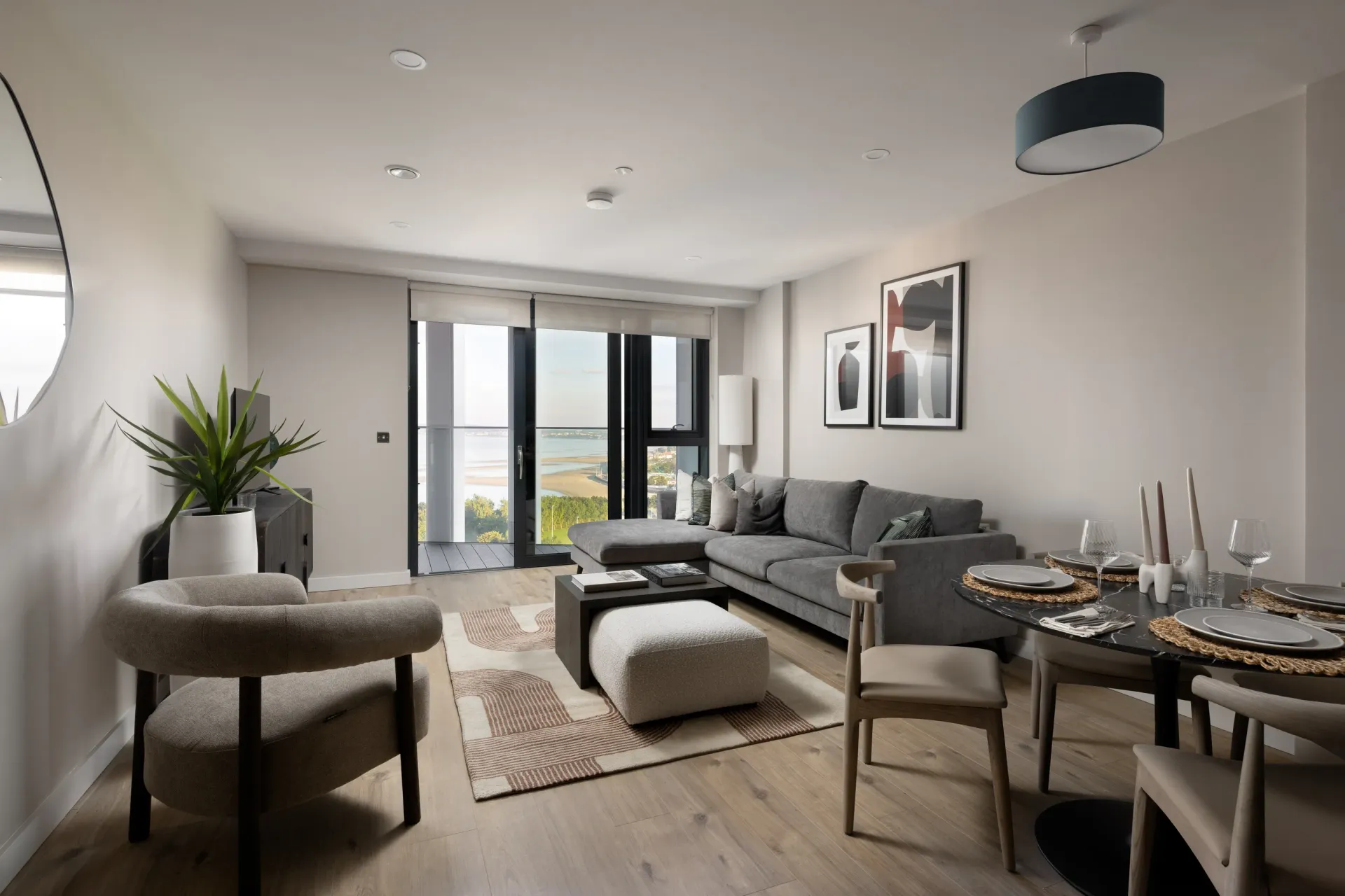 Bright modern living room with gray sofa, dining table, balcony doors, and indoor plants