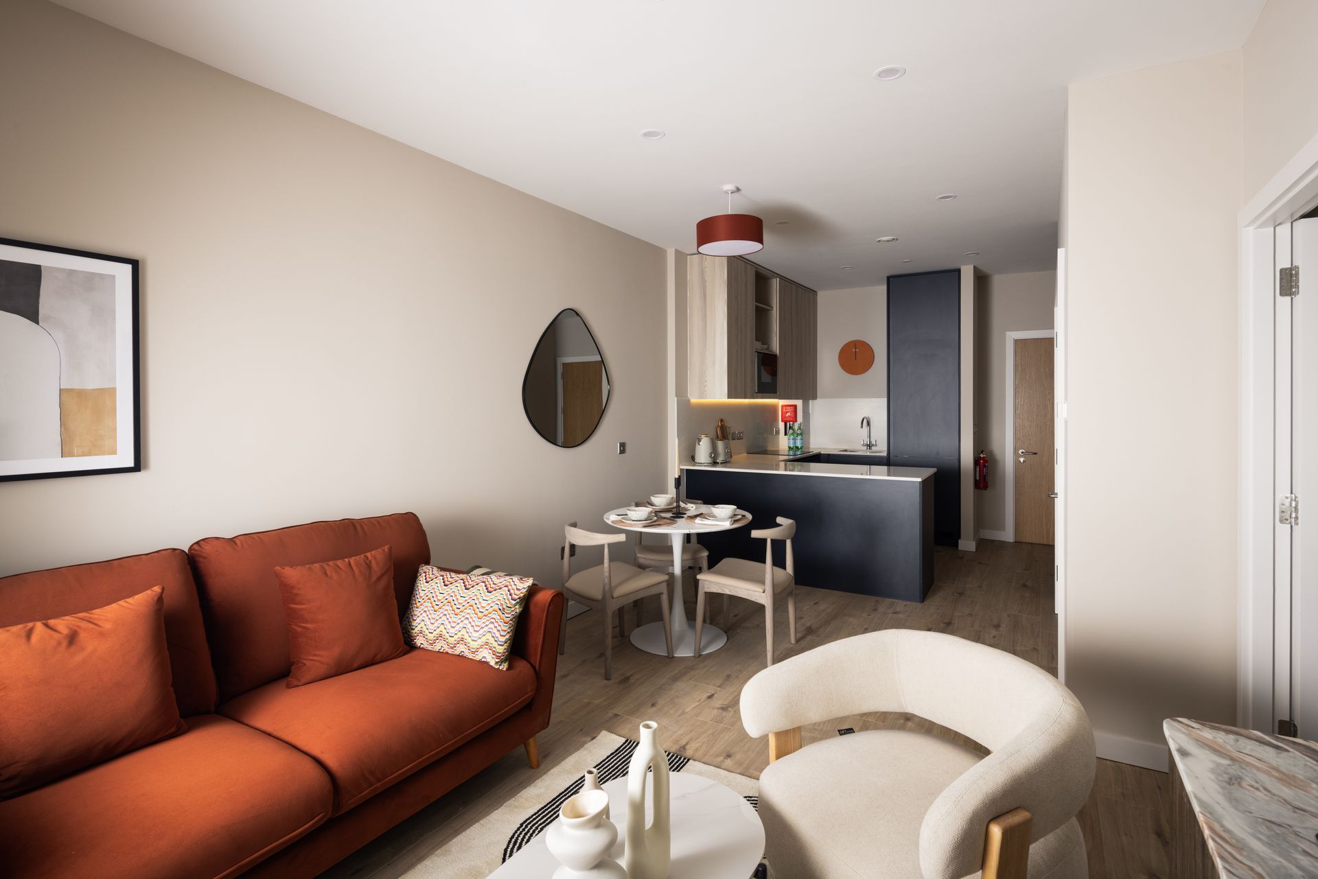 Modern living room with orange sofa, cream chair, small dining table, and open kitchen nook.