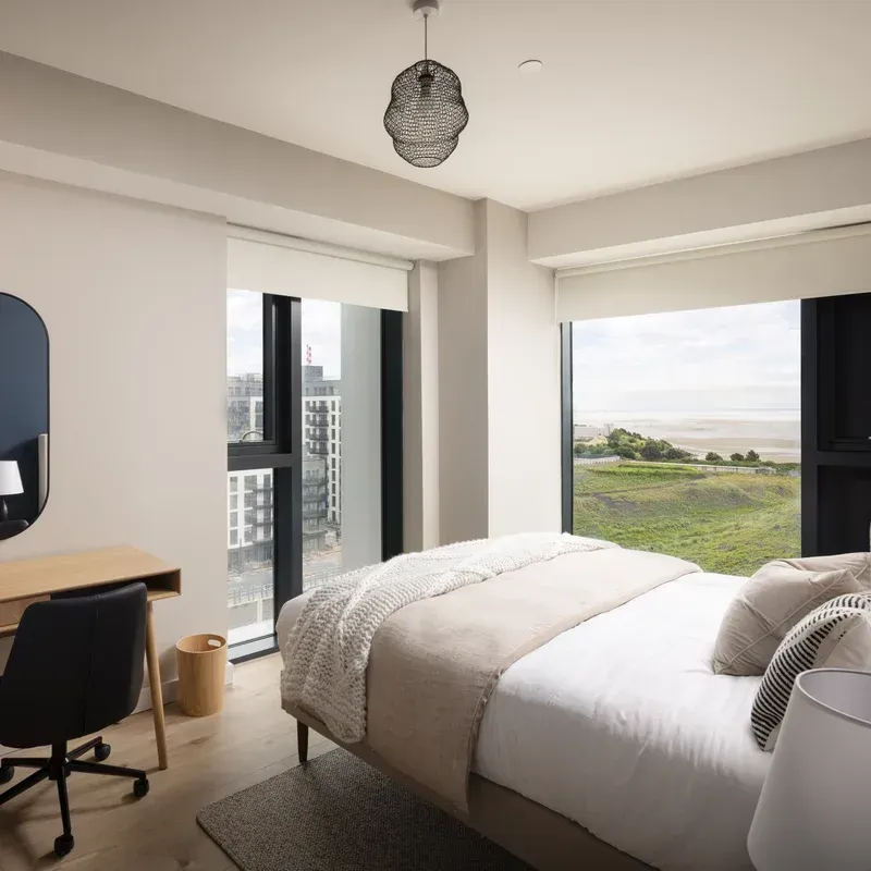 Modern bedroom with a bed, desk, and large windows overlooking green fields and city buildings