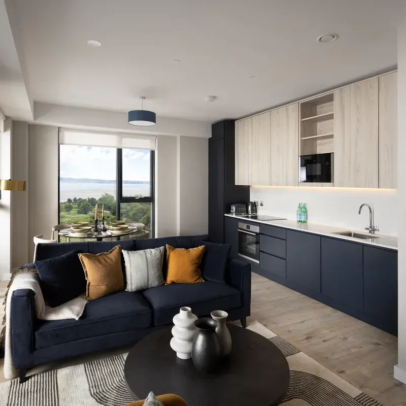 Modern open-plan living room with navy sofa, round coffee table, and light wood kitchen by a large window