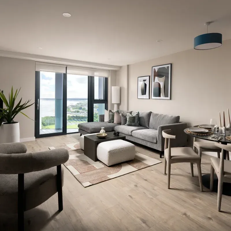 Modern living room with gray sofa, armchair, coffee table, dining nook, and large windows overlooking water.