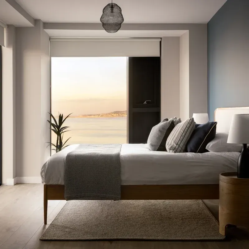 Modern bedroom with gray bedding, open window, and ocean view at sunset