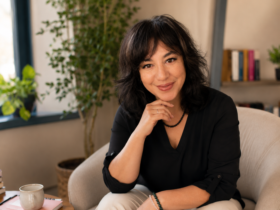 Een vrouw in een zwarte top glimlacht aan een tafel in een lichte kamer met planten en boeken.