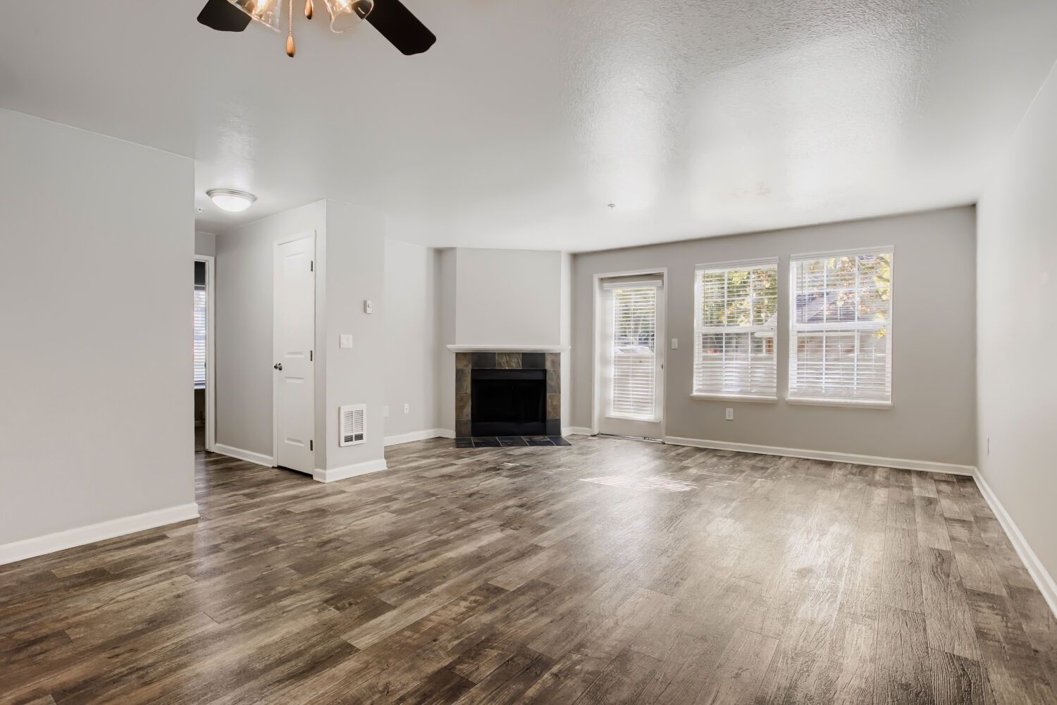 Empty living room with gray walls, wood-look floors, fireplace, and large windows letting in light