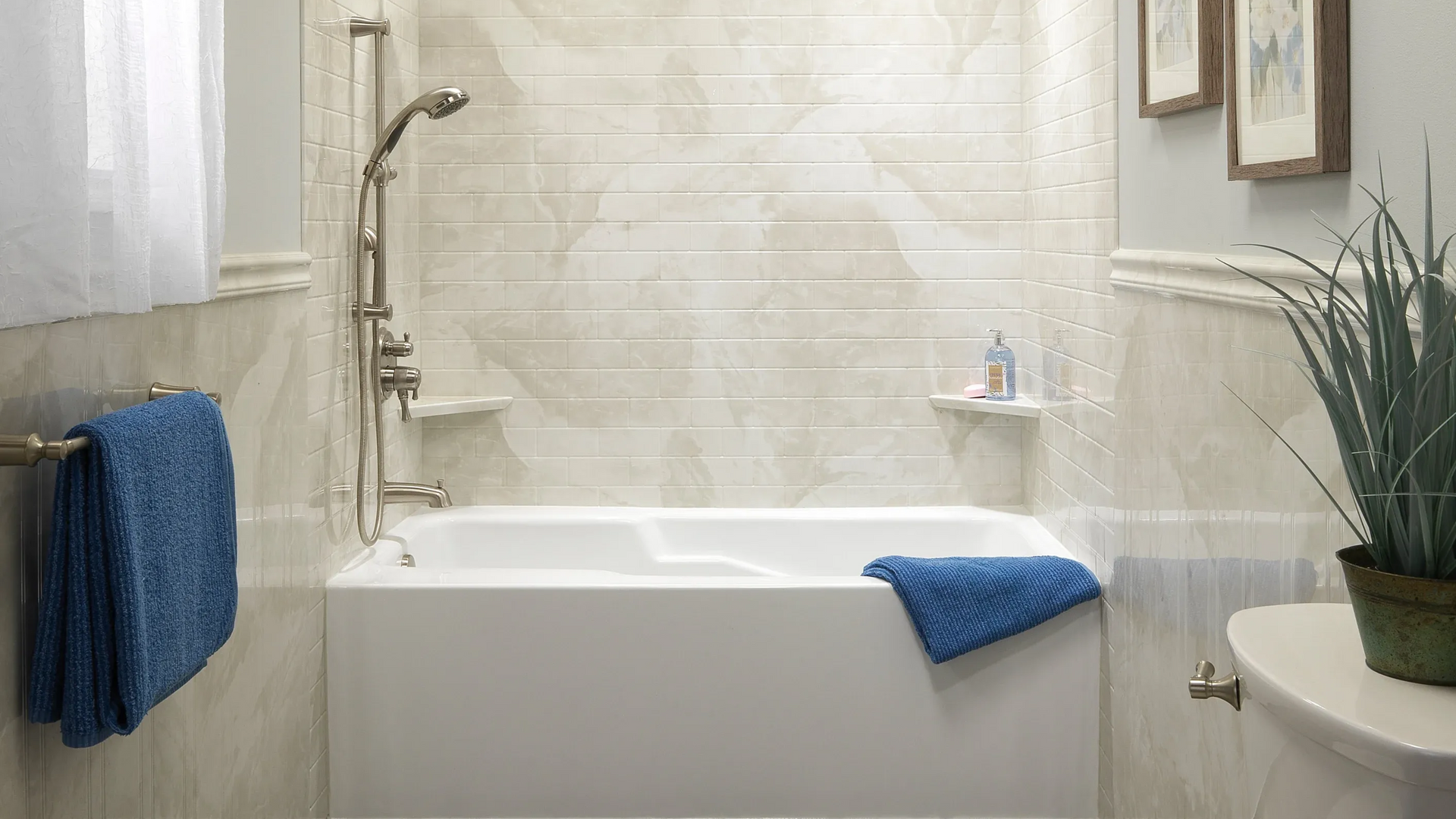 Bright bathroom with white bathtub, tiled shower wall, blue towels, and a potted plant.
