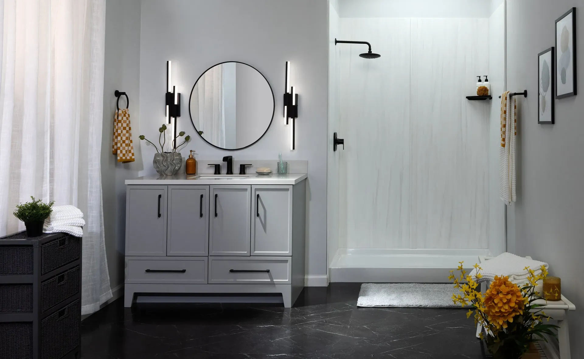 Modern gray bathroom vanity with round mirror, black fixtures, white shower, and flowers on the floor.