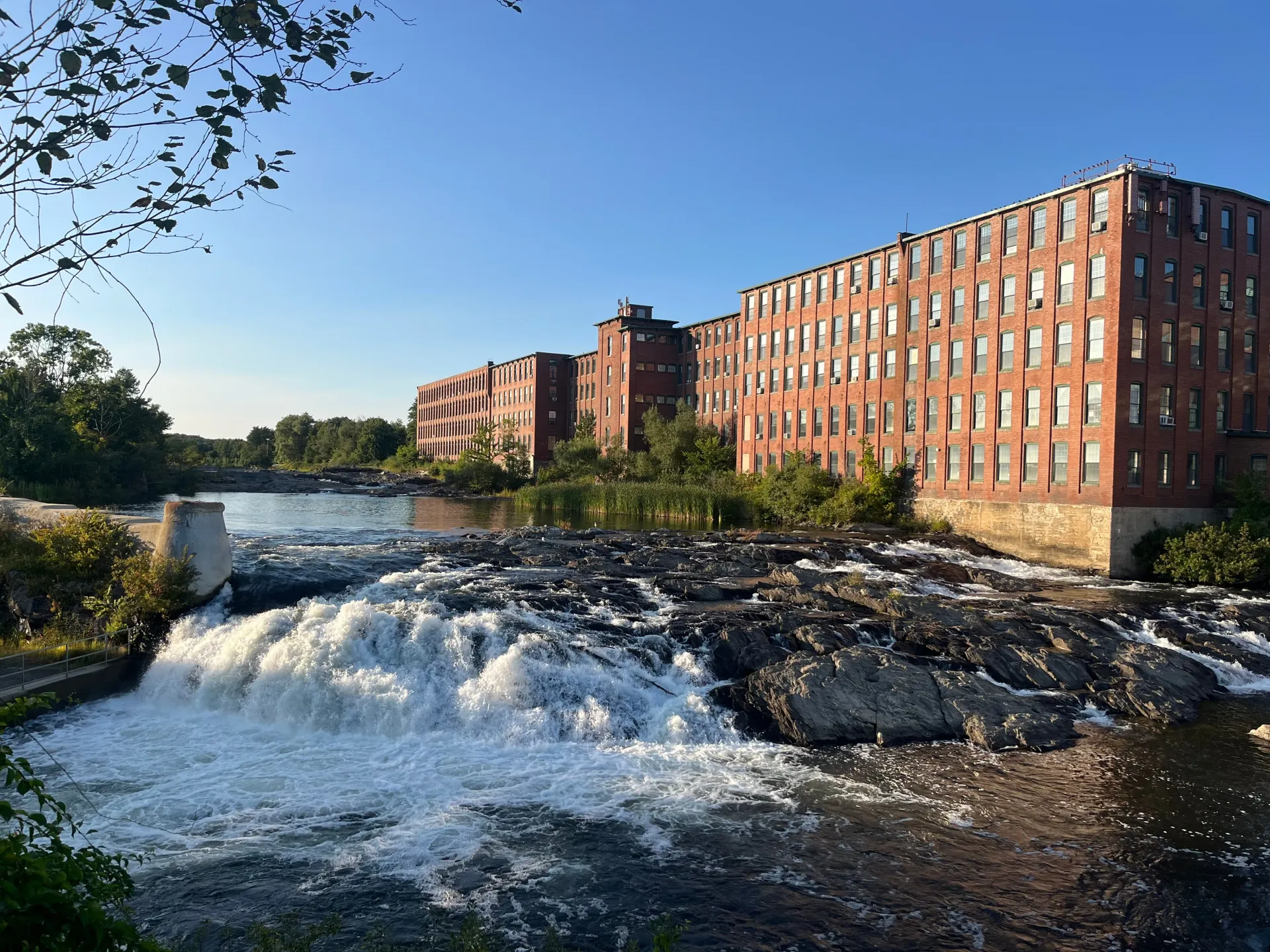 River waterfall beside red brick mill buildings under a clear blue sky
