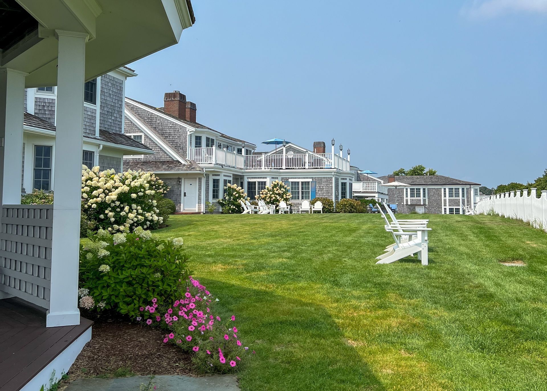A charming coastal home with gray shingles, vibrant flowers, Adirondack chairs on a lush green lawn, and a sunny patio adorned with white railings under a clear blue sky.