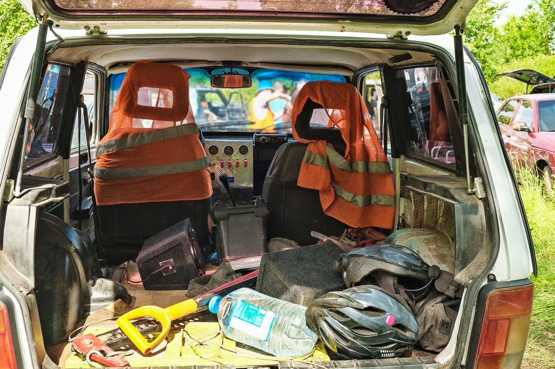 Open trunk of a white car, filled with gear. Orange safety vests cover seats. Shovel, water bottle, and helmets visible.