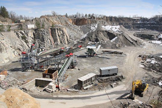 Quarry with conveyor belts, heavy machinery, and piles of rocks under a blue sky.