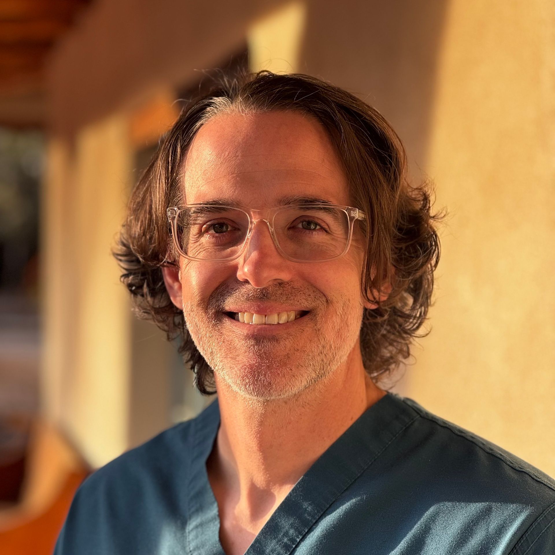 Smiling person in glasses and a blue shirt, standing in warm sunlight indoors.