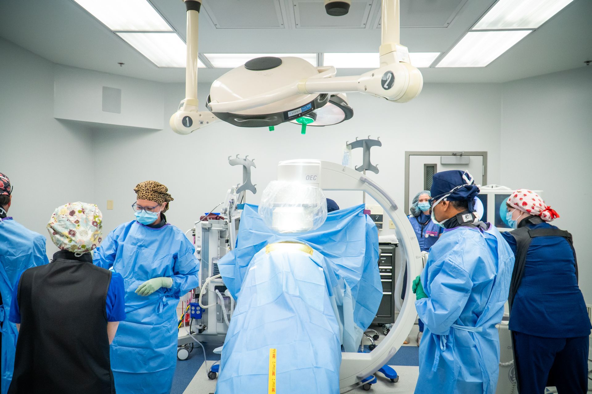 Surgical team in blue gowns around an operating table under bright OR lights and ceiling-mounted equipment