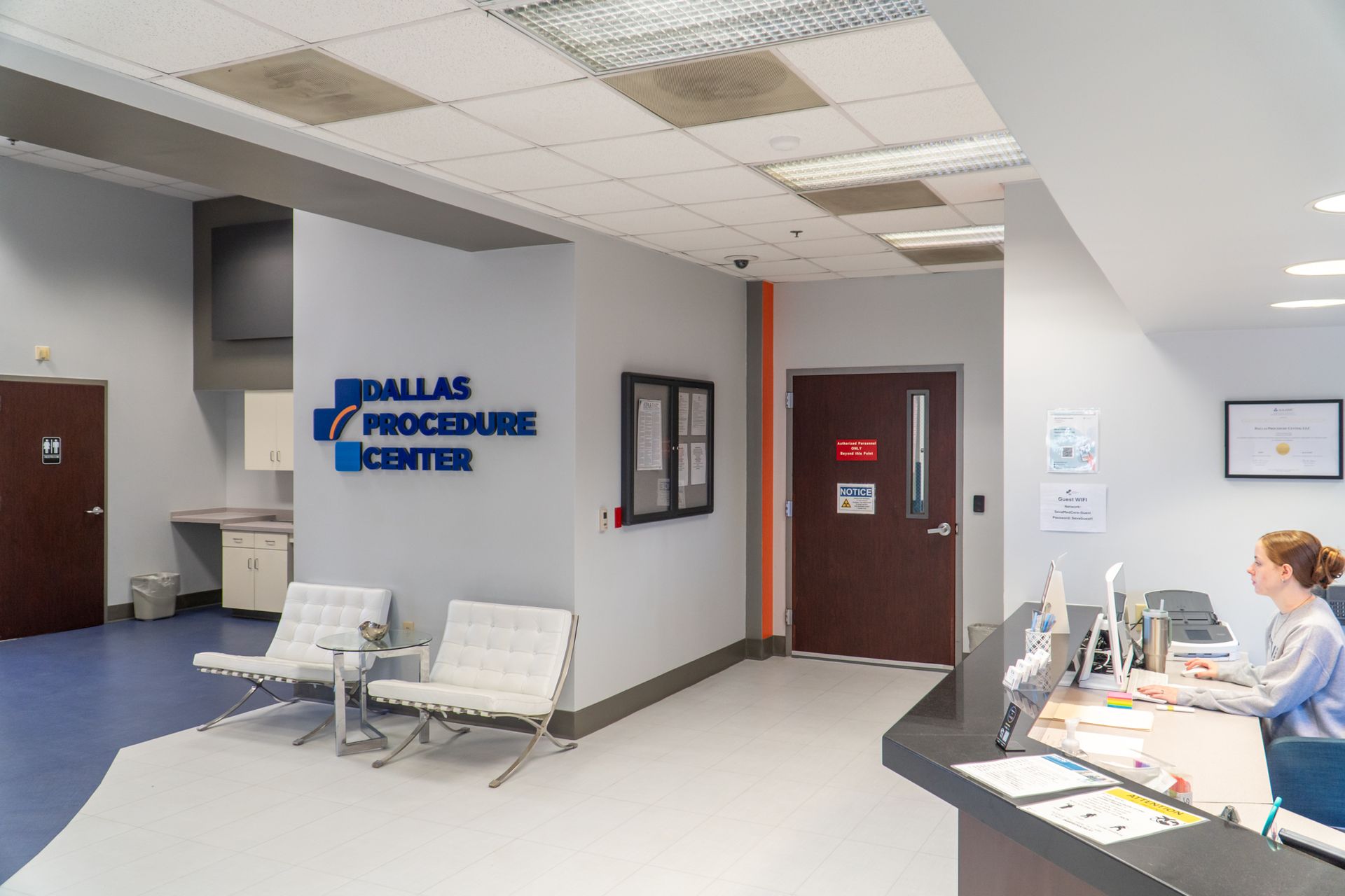 Clinic waiting area with blue chairs, reception desk, and Dallas procedure center sign.
