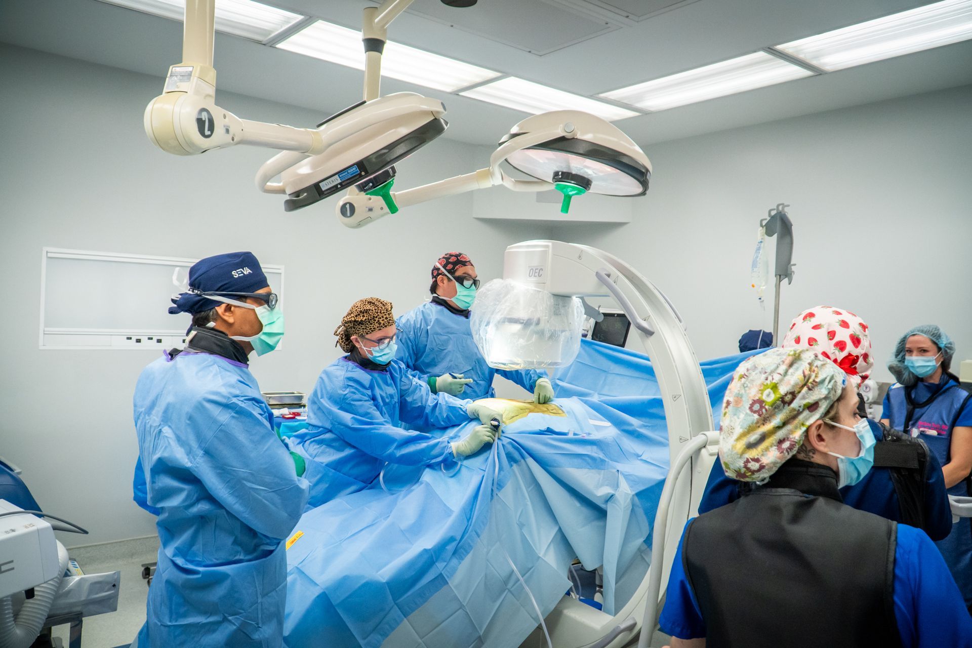 Surgeons in blue scrubs perform an operation in a bright operating room under overhead surgical lights