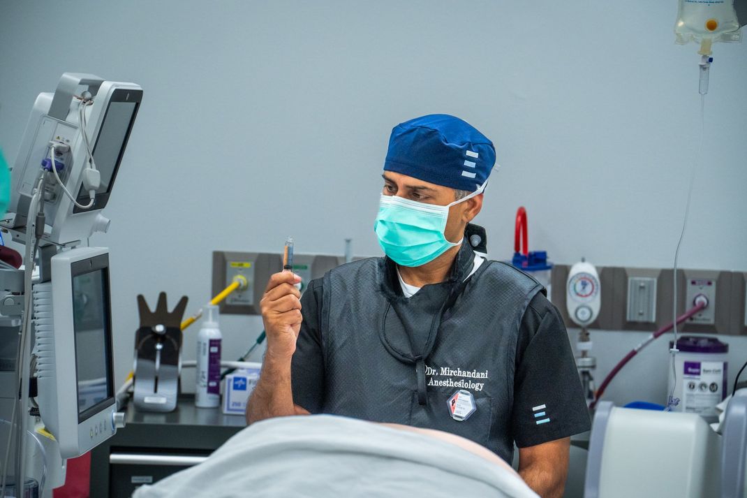 Masked dentist in scrubs beside a dental chair, holding up a gloved hand in a clinic.