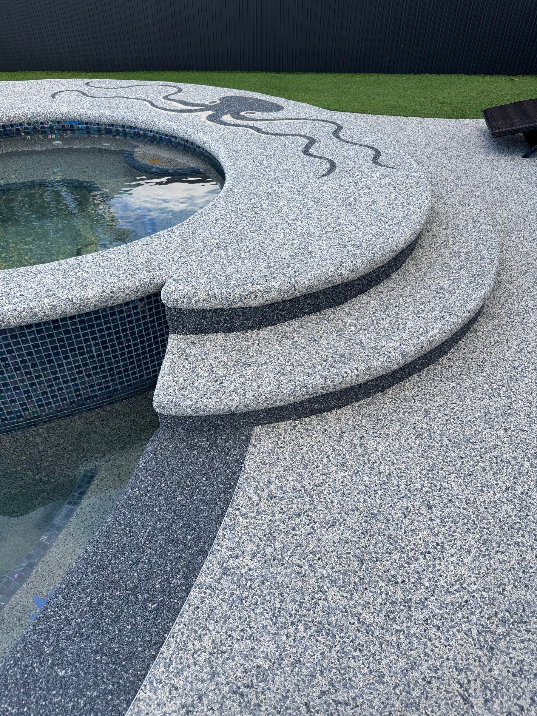 Curved stone steps beside a tiled pool with gray speckled paving and grass beyond