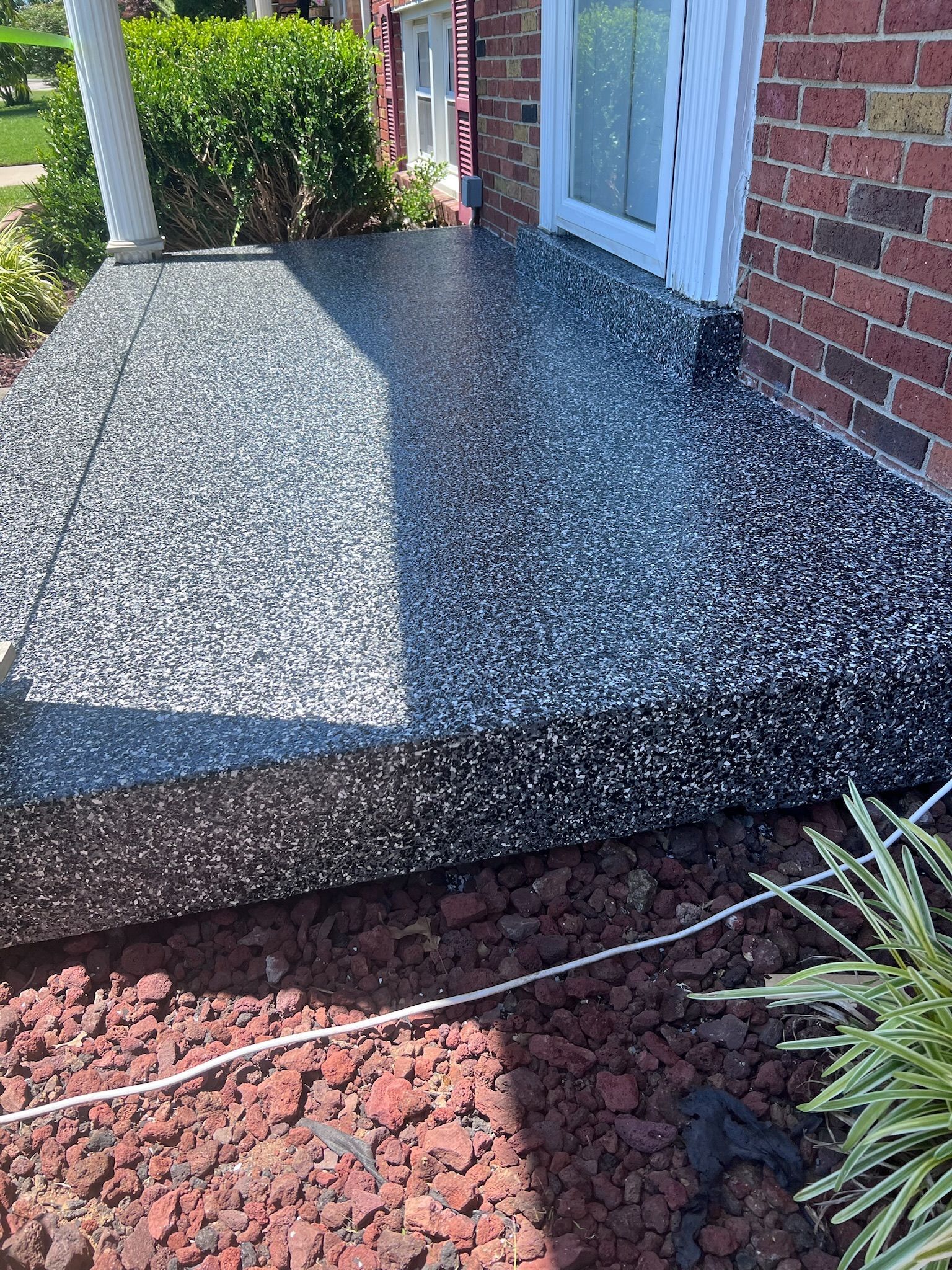 Black and gray speckled concrete porch. Red brick house with a white door. Landscaping in the foreground.